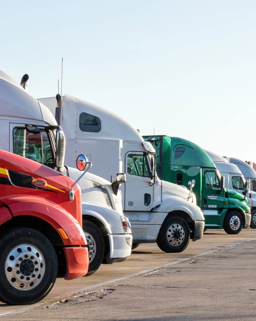 A line of parked semi trucks — a red Peterbilt, white sleepers and a green Heartland Express tractor — at sunrise